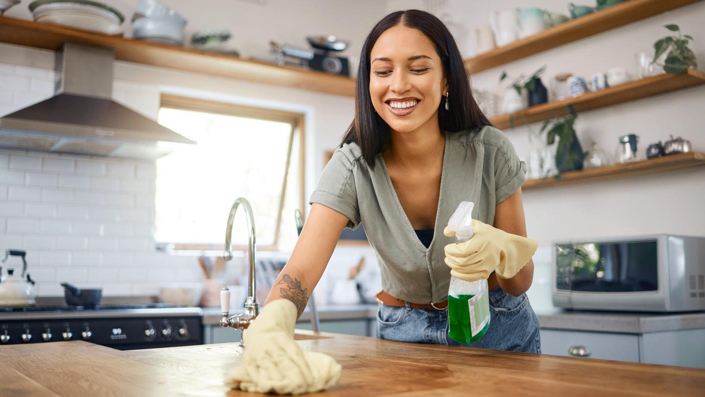 One smiling young mixed race woman wearing protective gloves and using a spray bottle with sanitiser to wipe her kitchen counters at home. Hispanic cleaning, staying healthy and hygienic with chores